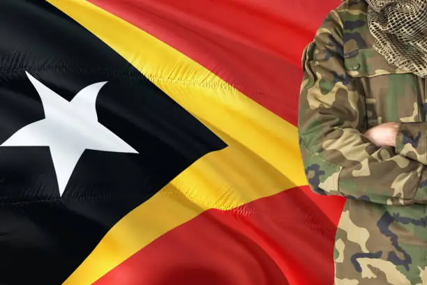 Photo of Crossed arms Timorese soldier with national waving flag on background - East Timor Military theme.