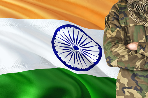 Crossed arms Indian soldier with national waving flag on background - India Military theme.