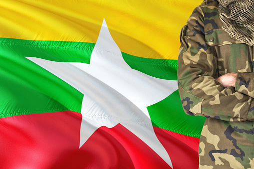 Crossed arms Burmese soldier with national waving flag on background - Burma Military theme.