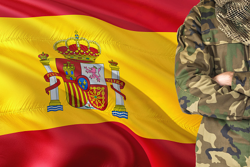 Crossed arms Spanish soldier with national waving flag on background - Spain Military theme.