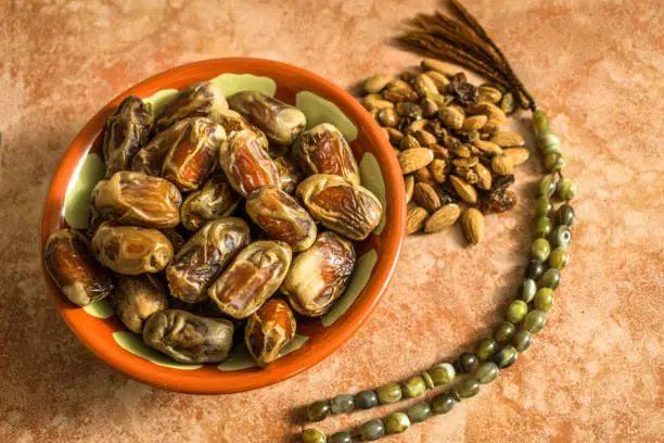 lots of fresh and healthy dates kept in a muddy Arabian traditional pot and almonds and a religious Tasbeeh  kept of a marble table and ready to break fast in the holy month of Ramadan