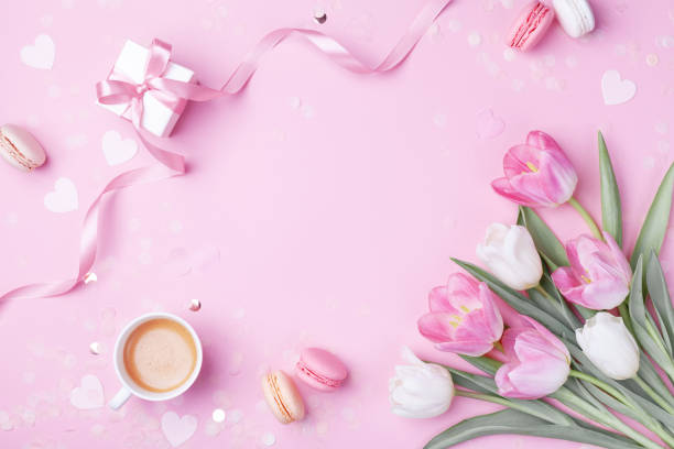 Photo of Morning cup of coffee, cake macaron, gift or present box and spring tulip flowers on pink background. Beautiful breakfast for Women day, Mother day. Flat lay.
