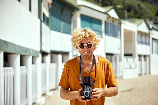 Portrait of smiling mid adult man with vintage camera. Male hipster is standing against beach huts. He is enjoying summer at beach.