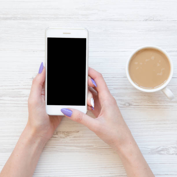 istock Female hands using smartphone and drinking latte over white wooden table, top view. Flat lay, from above. 1132058078