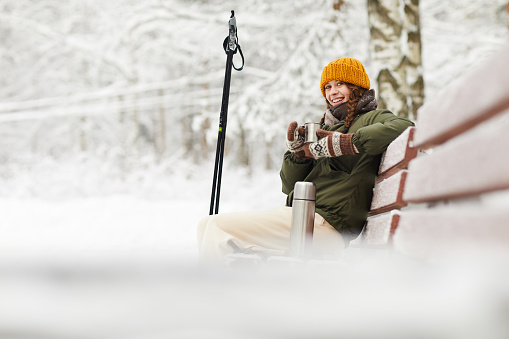 Side view portrait of active young woman smiling at camera while enjoying hot cocoa sitting on bench in beautiful winter forest, copy space