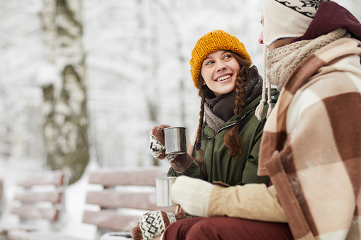 Side view portrait of loving young couple wrapped in blanket drinking hot cocoa outdoors sitting on bench beautiful winter forest, copy space