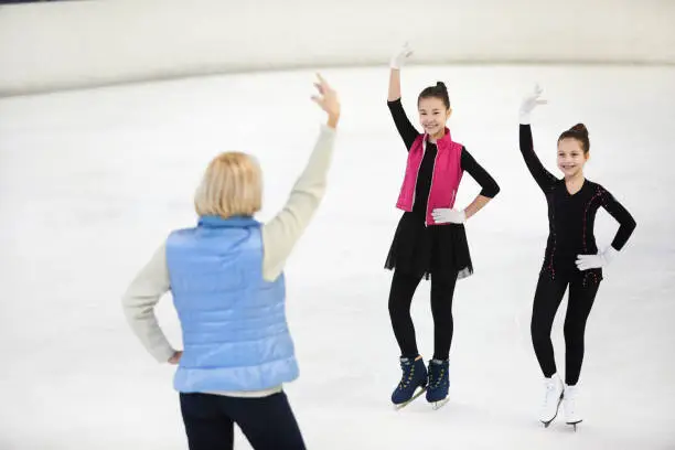 Full length portrait of two happy girls figure skating on rink with female coach giving instructions, copy space