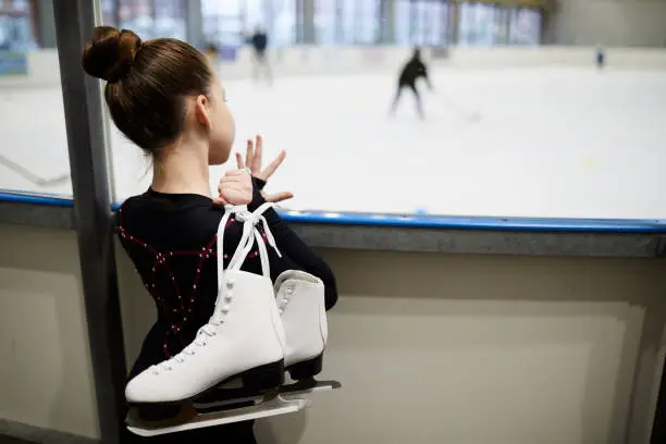 Back view portrait of future figure skater standing by ice rink and watching training, copy space