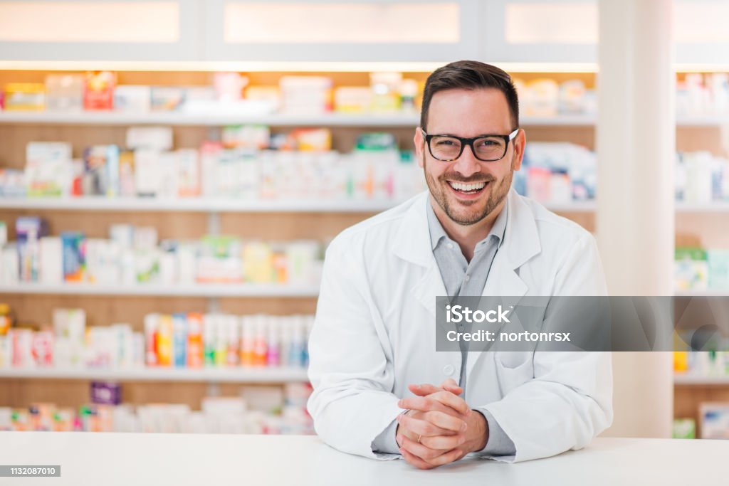 Retrato de un joven farmacéutico alegre apoyado en un mostrador en la farmacia, mirando a la cámara. - Foto de stock de Farmacéutico libre de derechos