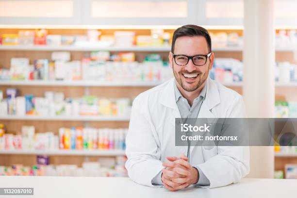 Retrato De Un Joven Farmacéutico Alegre Apoyado En Un Mostrador En La Farmacia Mirando A La Cámara Foto de stock y más banco de imágenes de Farmacéutico