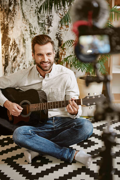 istock Online tutorial. Young music blogger playing the guitar and smiling, while recording online lesson for his subscribers 1132090516