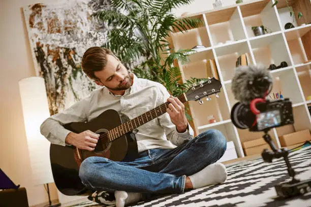 Photo of Learning guitar. Bearded male music blogger sitting on the floor at home and recording new guitar lesson for Youtube channel.