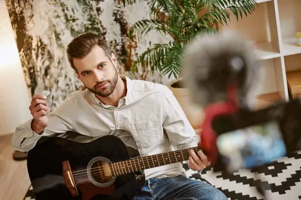 Photo of Learning guitar. Close up of attractive male music blogger sitting on the floor and showing how to play guitar with camera on foreground
