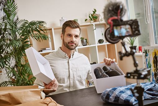 Fashion blog. Smiling male blogger opening box with sneakers while recording new video at home on a digital camera. Fashion blog. Beauty. Video review.