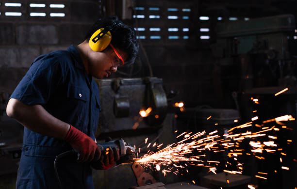 Mechanical engineer worker wearing safety equipment and operating a angle grinder on his workbench with metal held in an iron vice in a low light workshop with flash sparks stock photo
