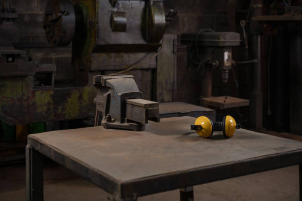 Industrial mechanical cast iron vice bolted to a metal work station table in a dark traditional old style repair workshop next to yellow ear defenders stock photo
