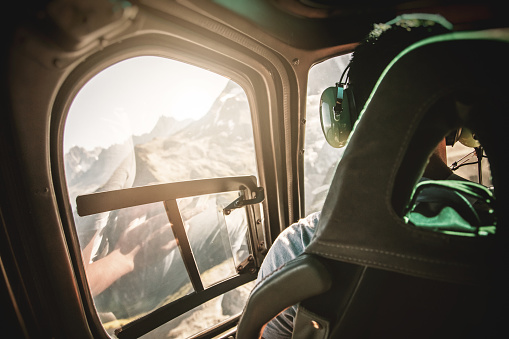 Helicopter cockpit interior with rear view of unrecognizable men co-pilot, flying high up altitude over Mont Blanc massif in French Alps mountains at sunset. The men is unrecognizable and looking through window the amazing nature landscape. All brands from flying instruments were removed.