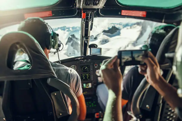 Interior of helicopter cockpit with flying instruments (brands were removed) and with unrecognizable pilot and co-pilot, and blurred tourist's arms taking photos with a mobile phone of the flight over Mont Blanc massif in French Alps mountains. Taken in Haute-Savoie, Auvergne-Rhone-Alpes region in France (Europe).