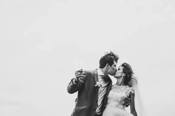 Photo of Romantic couple newlyweds, bride and groom is kissing on the background of a blue sky in park. Peaceful romantic and joyful wedding moment on nature in the field or water. Black and white photo.