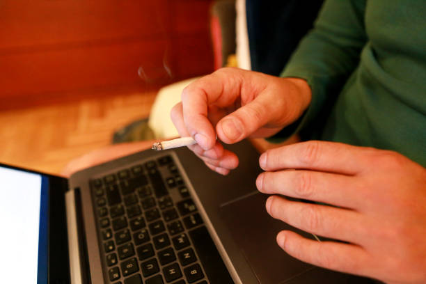 istock Man working on a laptop at home office, hand typing on keyboard with a cigarette in his hand is holding and smoking, close up. Businessman using notebook computer for working on his lap. Top view. 1132119364