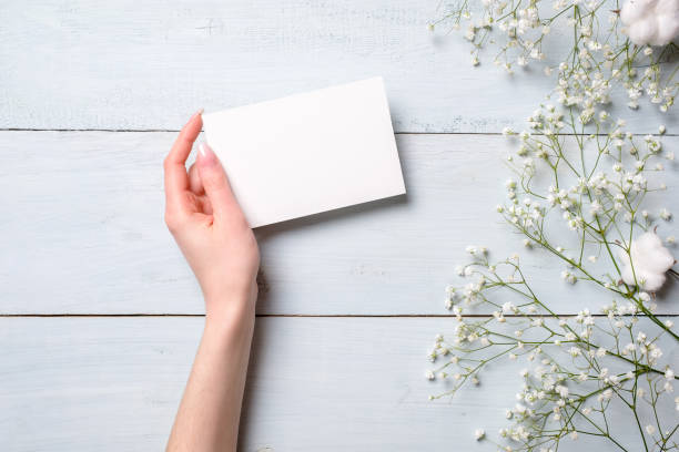 Womens hand holding blank paper card on light blue wooden background. Rustic table with flowers, wedding invitation card, letter, greeting card for Womens day. Trendy minimal design, Flat composition
