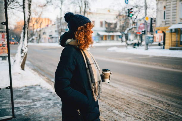 Young pretty woman wearing knitted hat drinking coffee and waiting for bus at stop on cold sunny winter day.