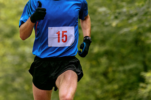 athlete runner in blue t-shirt with number running on background green trees