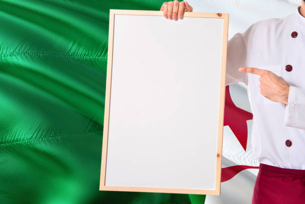 Algerian Chef holding blank whiteboard menu on Algeria flag background. Cook wearing uniform pointing space for text.