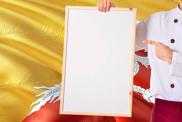 Bhutanese Chef holding blank whiteboard menu on Bhutan flag background. Cook wearing uniform pointing space for text.