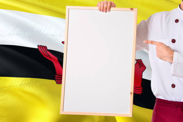 Bruneian Chef holding blank whiteboard menu on Brunei flag background. Cook wearing uniform pointing space for text.