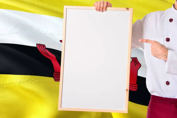 Photo of Bruneian Chef holding blank whiteboard menu on Brunei flag background. Cook wearing uniform pointing space for text.