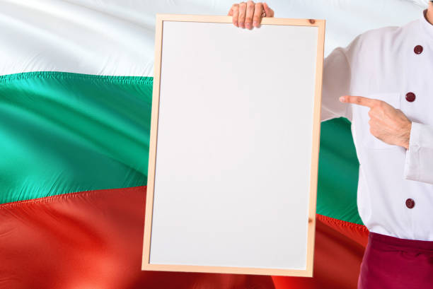 Bulgarian Chef holding blank whiteboard menu on Bulgaria flag background. Cook wearing uniform pointing space for text.