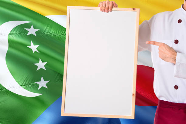 Comoran Chef holding blank whiteboard menu on Comoros flag background. Cook wearing uniform pointing space for text.