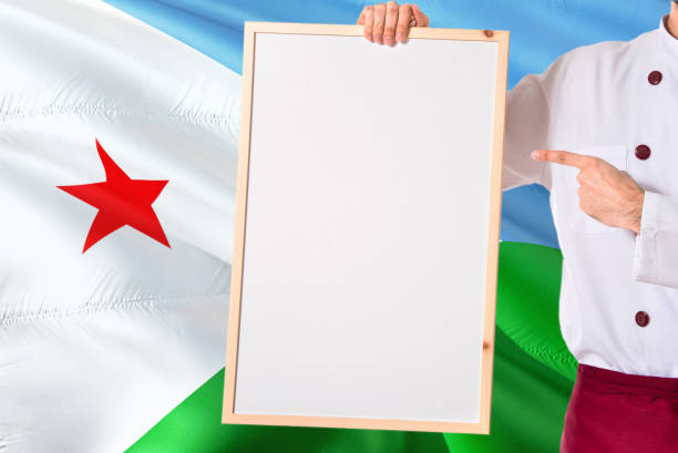 Chef holding blank whiteboard menu on Djibouti flag background. Cook wearing uniform pointing space for text.