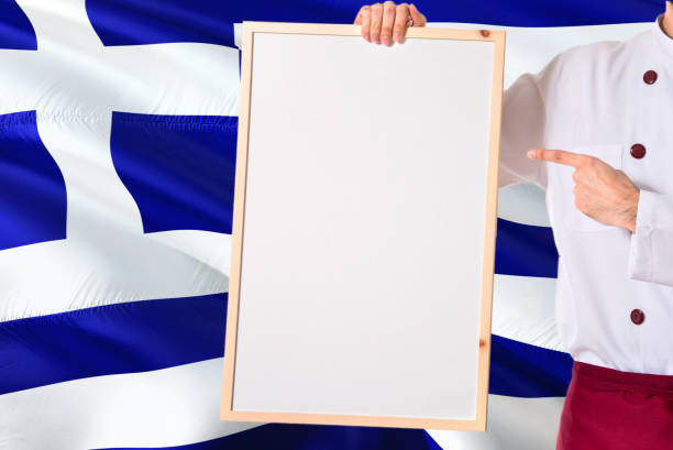 Greek Chef holding blank whiteboard menu on Greece flag background. Cook wearing uniform pointing space for text.