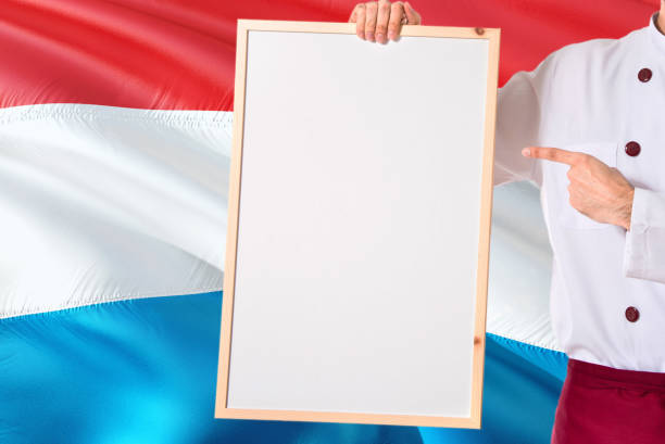 Chef holding blank whiteboard menu on Luxembourg flag background. Cook wearing uniform pointing space for text.
