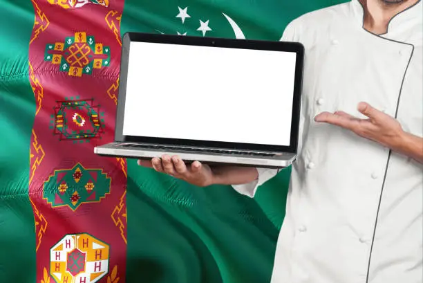 Photo of Turkmen Chef holding laptop with blank screen on Turkmenistan flag background. Cook wearing uniform and pointing laptop for copy space.