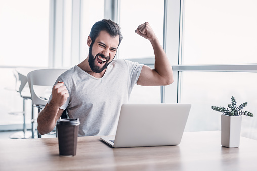 In the office businessman sitting at the desk, using laptop finishes project and wins big. Makes successful gestures, raises arms in celebration. His cup of coffee is in front of him