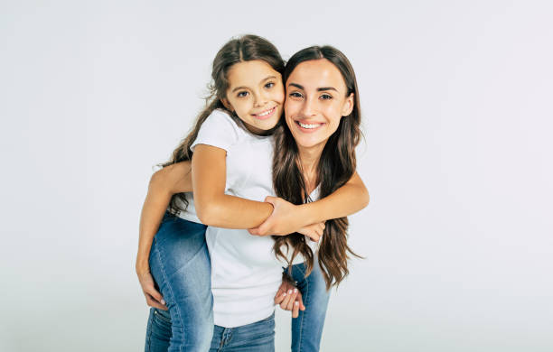 beautiful young brunette mother holds on back her cute little daughter in white t-shirts isolated in studio - family child portrait little girls imagens e fotografias de stock