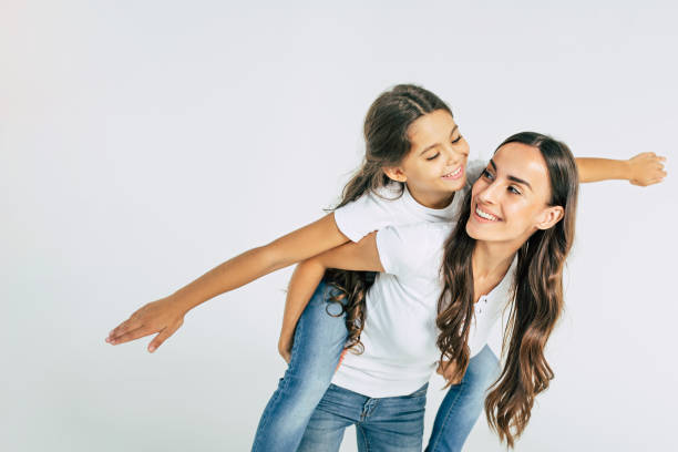 schöne junge brünette mutter hält auf dem rücken ihre niedliche kleine tochter in weißen t-shirts im studio isoliert - family child portrait little girls stock-fotos und bilder