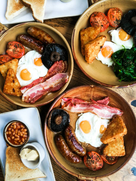 Traditional Full English Fried Breakfast Overhead color image depicting several plates of freshly cooked full English fried breakfast. The traditional meal consists of sausages, rashers of bacon, baked beans, toasted bread, mushrooms, hash browns, fried eggs and grilled tomatoes. Room for copy space. english breakfast stock pictures, royalty-free photos & images