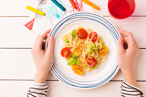 Kid's meal - spaghetti with cherry tomatoes and basil. Colorful dinner - plate in child's hands on white wooden table. Plate captured from above (top view, flat lay).