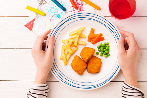 Kid's meal - chicken nuggets, french fries, carrot and green peas. Colorful dinner - plate in child's hands on white wooden table. Plate captured from above (top view, flat lay).