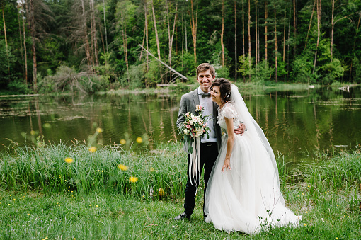 Portrait of an attractive groom and bride standing on nature in the park. Happy and joyful moment. Romantic couple of newlyweds near pond. Wedding ceremony near lake. Photography. Photo.