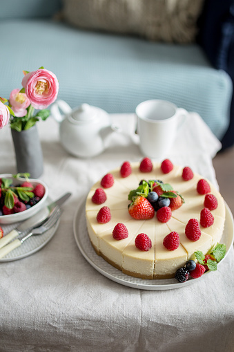 Cheesecake, homemade cake with strawberry berries in a gray plate, with cup, teapot, fork, knife, fresh berries, vase with flowers, side view. Sweets on table in living room. Organic product