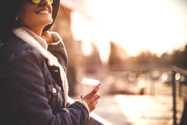 Vertical portrait of a cute urban hipster woman holding a pumpkin spice coffee drink. She is sitting outside of a coffee shop relaxing and enjoying her beverage. She is dressed for fall season fashion wearing a cute neck scarf and black jacket.