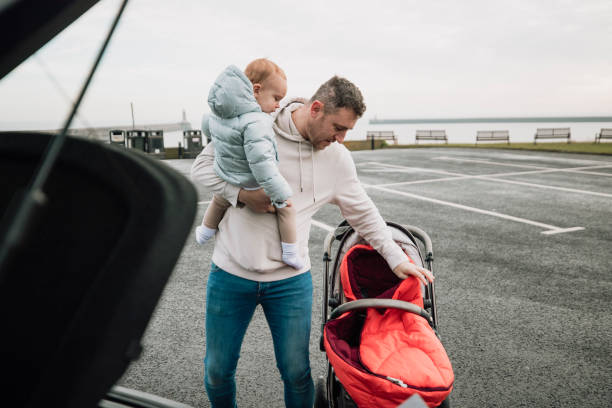 Preparing the Pushchair in the Parking Lot Multi-tasking father is in a parking lot, holding his baby boy in one arm while setting up his pushchair with the other. northern europe family car stock pictures, royalty-free photos & images