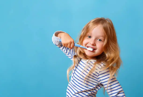 A little girl in striped pajamas is brushing her teeth with a toothbrush. The concept of daily hygiene. On a blue background