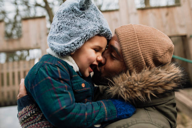 Dad and his son in the park during winter time. He is holding his son in his arms and giving him a kiss on the cheek.