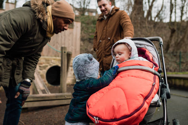 Play Date Two Dads are in the park with their baby sons during winter time. They are wearing warm clothing. One of the babies is greeting and smiling at the other baby. baby stroller winter stock pictures, royalty-free photos & images